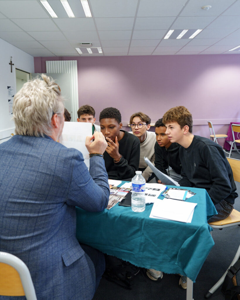 Cinq lycéens assis à la table d'un exposant, vu de dos, écoutent attentivement ; brochures et bouteille d'eau sur une nappe verte.
