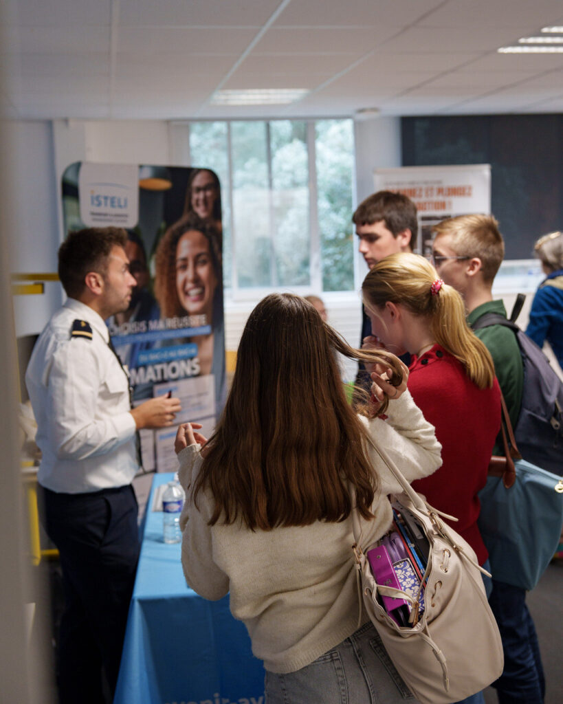 Une salle pleine de lycéens ; au stand de l'ISTELI, un intervenant en uniforme blanc répond à une élève.