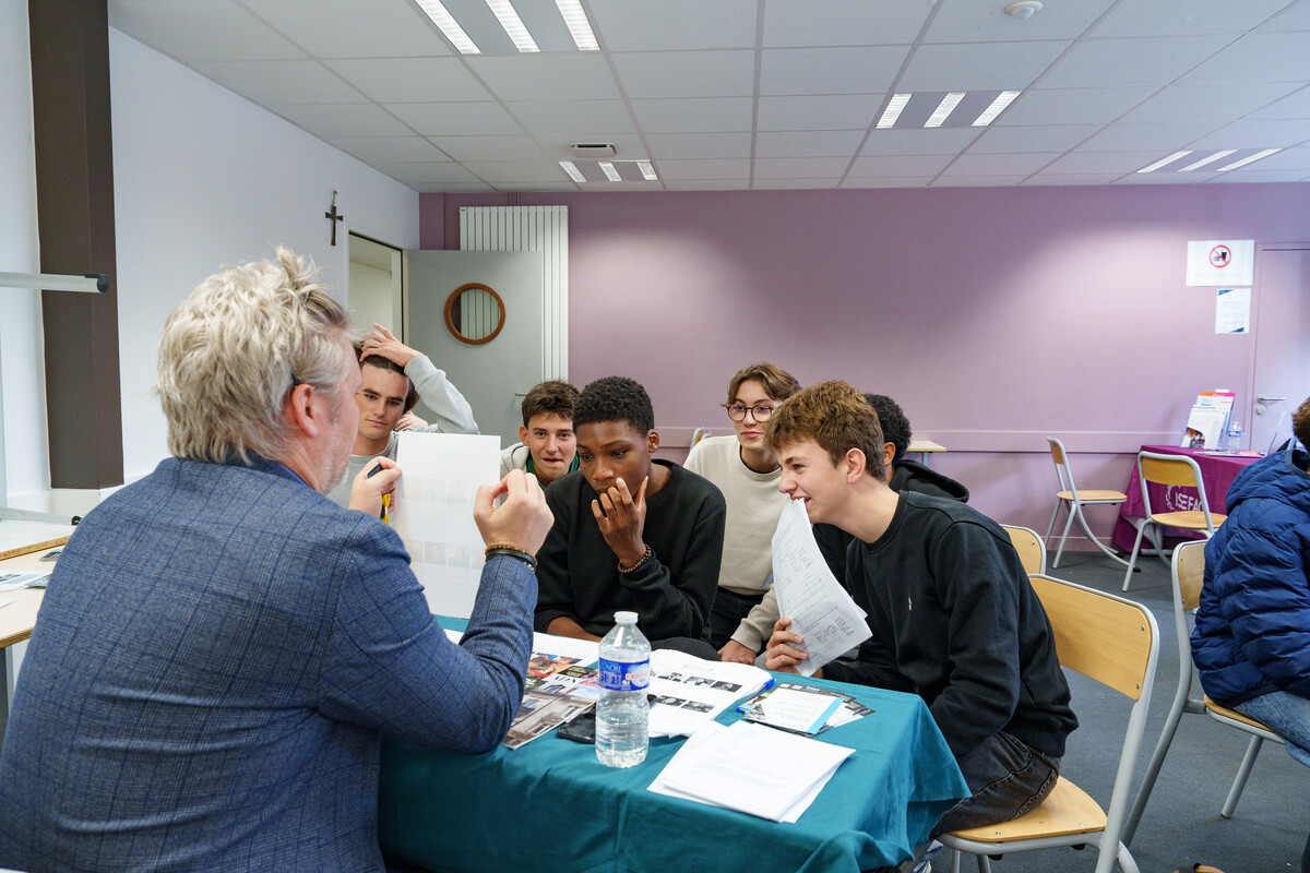 Des lycéens attentifs autour de la table d'un exposant, vu de dos, dans une salle du forum de l'orientation BDC Avenir.