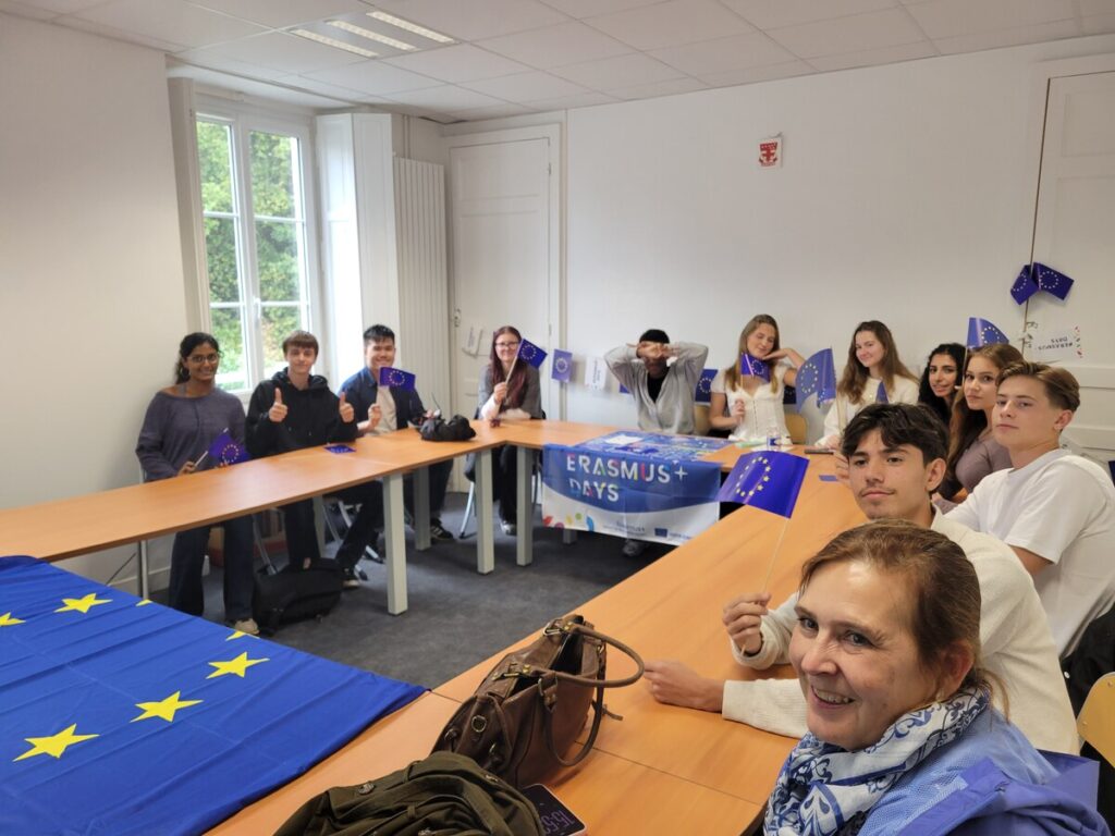 Dans une salle de classe aux couleurs de l'Europe, des élèves agitent de petits drapeaux européens autour d'une banderole « Erasmus+ Days » ; au premier plan, une femme sourit à l'objectif
