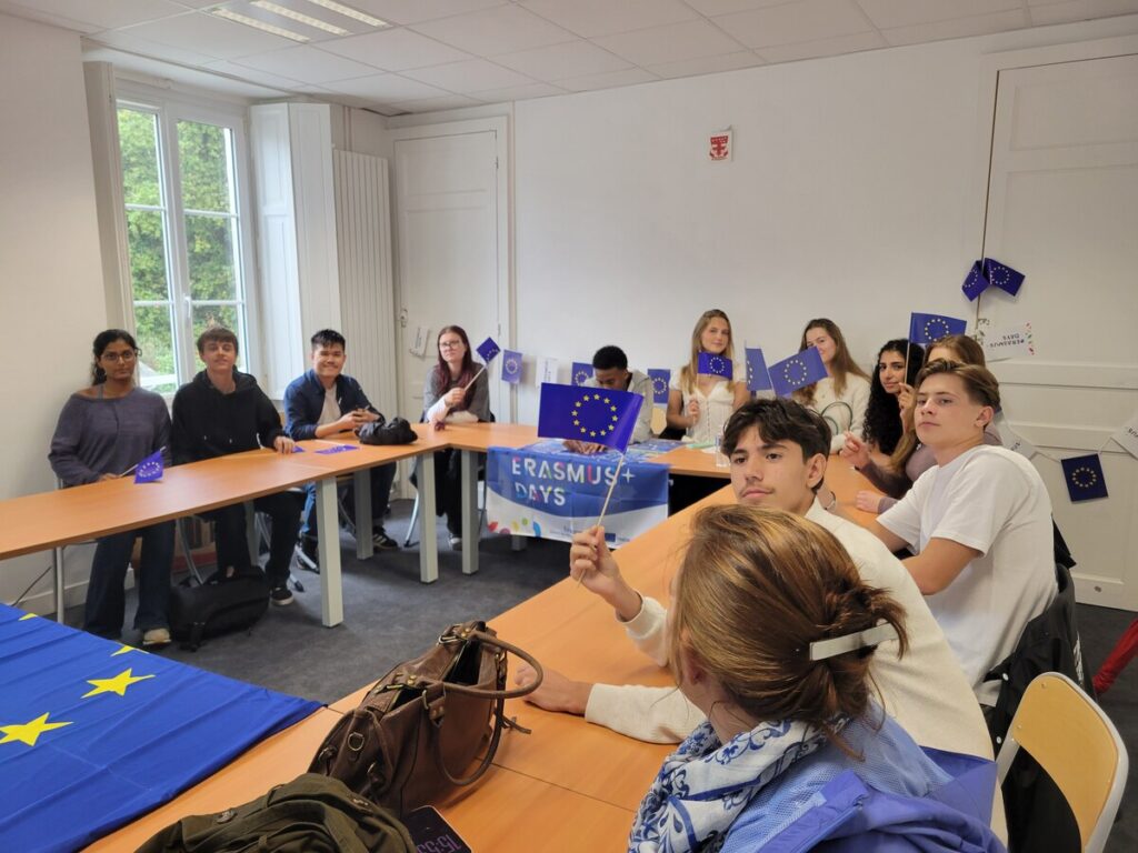 Autour de tables en U, des élèves agitent de petits drapeaux européens ; sur la table du fond, une banderole « Erasmus+ Days »