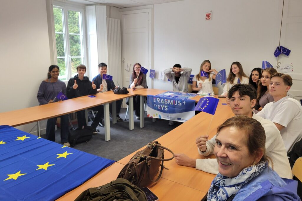 Dans une salle de classe aux couleurs de l'Europe, des élèves agitent de petits drapeaux européens autour d'une banderole « Erasmus+ Days » ; au premier plan, une femme sourit à l'objectif